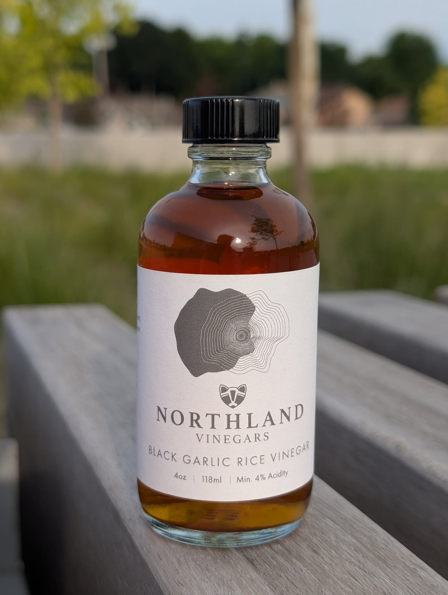 Bottle of Northland Vinegars Black Garlic Rice Vinegar on a wooden surface with a blurred outdoor background