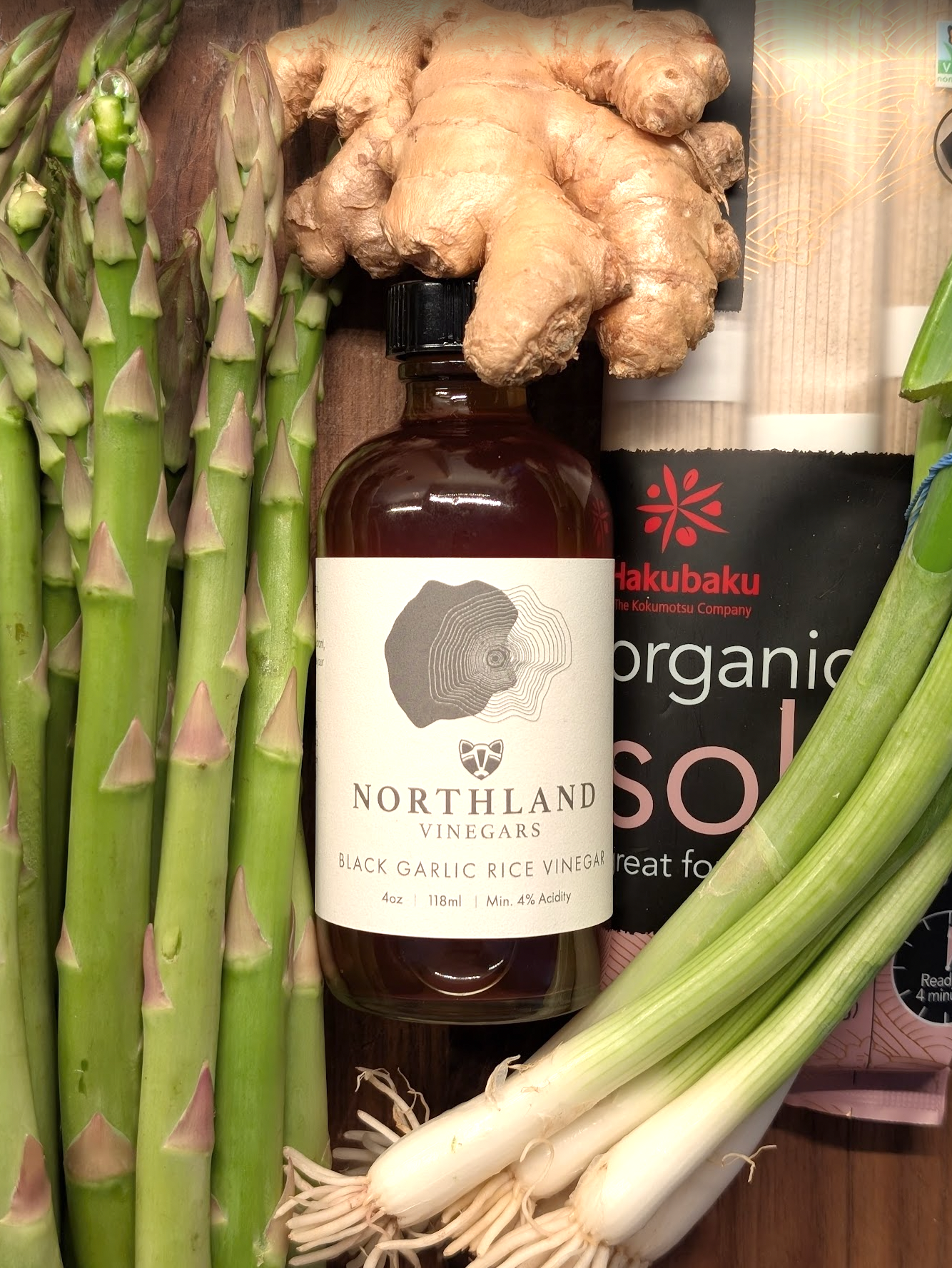 Bottle of Northland Vinegars black garlic rice vinegar surrounded by asparagus, ginger, and green onions on a wooden surface.