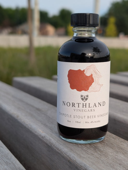 Bottle of Northland Vinegars chipotle stout beer vinegar on a wooden surface with a blurred outdoor background.